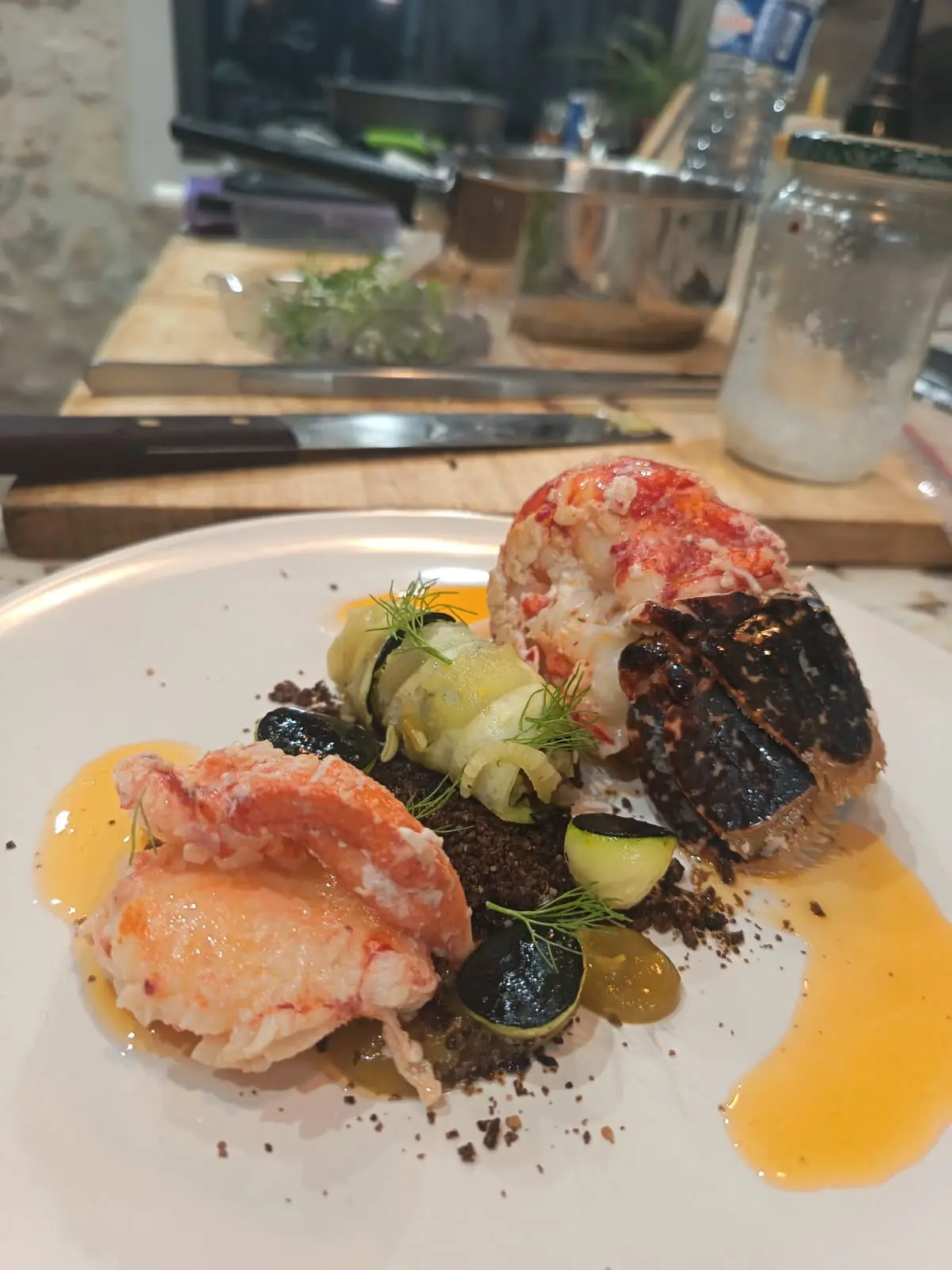 Queue et pince de homard, rouleau de courgette et sauce orange dans une assiette claire, avec une cuisine en arrière-plan