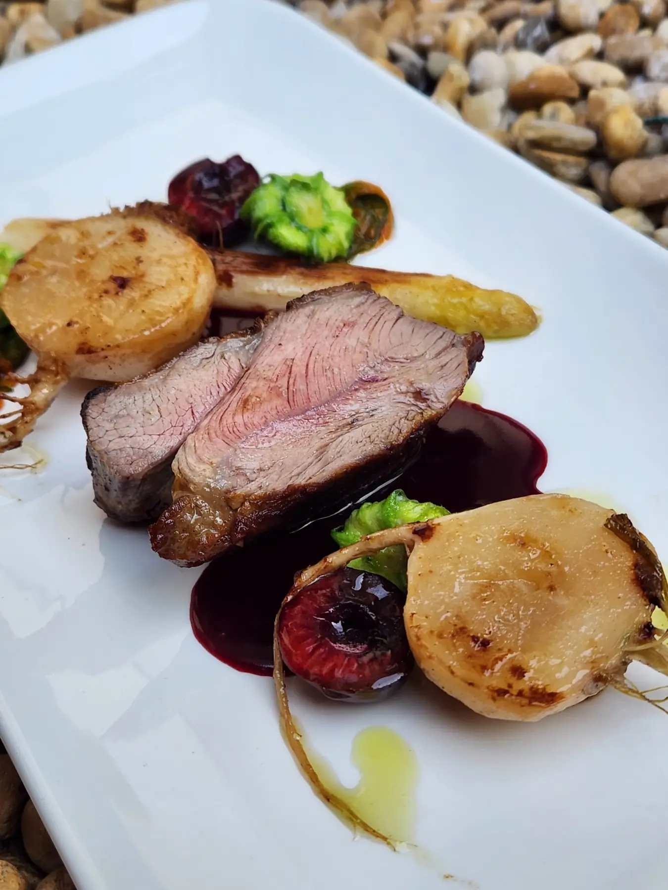 Bœuf rosé, pommes de terre et jus sombre dans une assiette rectangulaire blanche, posée sur des galets