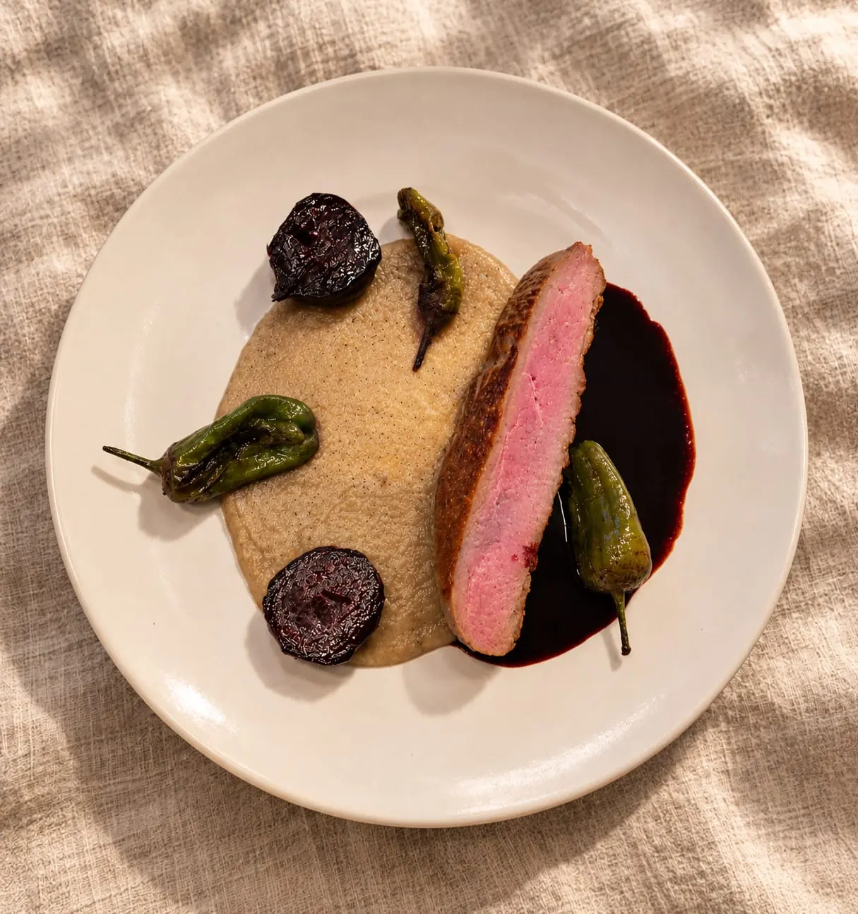 Une tranche de viande rosée sur une purée crème, avec des betteraves et des piments verts, dans une assiette crème posée sur du lin